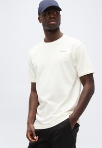 Άνδρας που φοράει απλό λευκό κοντομάνικο T-shirt με το κείμενο "30904" στο στήθος, μαύρο παντελόνι και μπλε καπέλο μπέιζμπολ, κοιτάζοντας προς τα πλάγια.