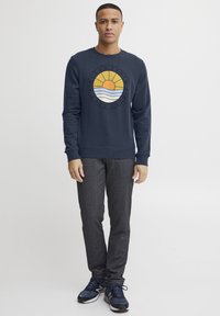 Tamsiai mėlynas džemperis su apvaliu grafiniu piešiniu, kuriame pavaizduoti saulė ir bangos. Po juo yra tekstas "BLEND DENIM ESTD 1993." Aprangą užbaigia pilkos kelnės ir tamsiai mėlyni sportbačiai.