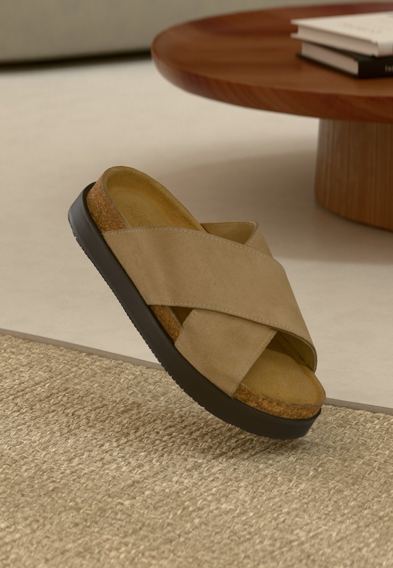 Beige keresztpántos szandál puha velúrból, parafa talpbetéttel és fekete gumi talppal, egy texturált szőtt felületen bemutatva.