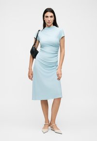 Γυναίκα με γαλάζιο μίντι φόρεμα με ρυτίδες, μπεζ μυτερά φλατ παπούτσια και μαύρη τσάντα ώμου, στέκεται μπροστά σε λευκό απλό φόντο.