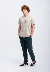 Beige katoenen T-shirt met een geometrische grafiek in blauw en roze, gecombineerd met donkergroene joggingsbroeken en zwarte Vans sneakers.
