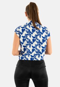 Blusa a maniche corte con un motivo floreale blu su sfondo bianco. Il top ha un colletto e un taglio corto, abbinato a pantaloni neri.