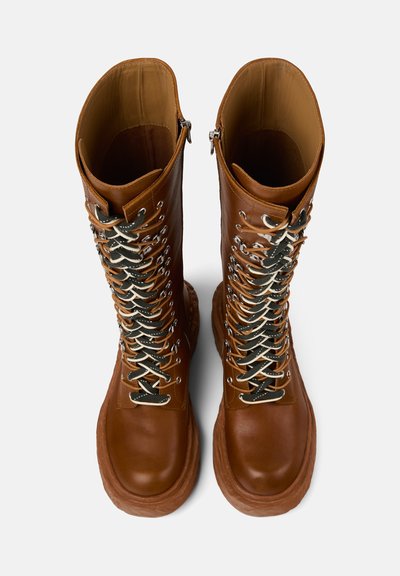 Botas de cuero marrón con cordones y caña alta, que presentan herrajes plateados, cordones verdes y crema, y una suela de goma texturizada.