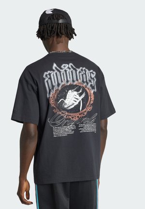 Mand iført sort Adidas-kasket og oversized sort T-shirt med Adidas-sko-grafik og tekst på ryggen, med ryggen mod kameraet.