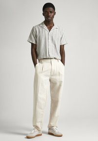 Kortärmad, randig skjorta i beige, ljusgrön och vit; löst sittande beige byxor; vita sneakers med grå accent och gummisulor.