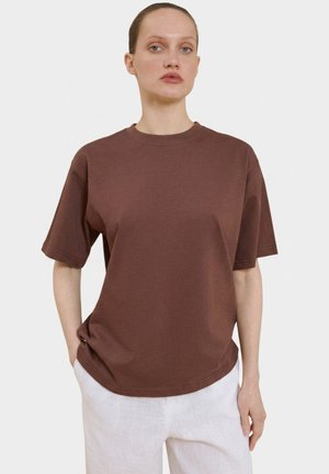 Kobieta o jasnej karnacji i związanych włosach, ubrana w jednolity brązowy t-shirt z krótkim rękawem i białe spodnie, stojąca z rękami w kieszeniach.