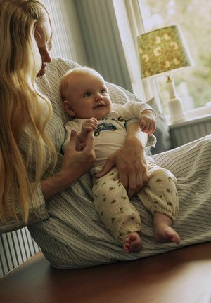 Baby iført en lysfarvet bodystocking med blå biltryk og mønstrede cremefarvede bukser med små designs, holdt af en voksen på en stribet overflade.