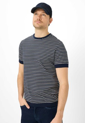 Mand med let skæg iført navyblå kasket, navyblå og hvide stribede kortærmede skjorte og mørke jeans, stående med den ene hånd i lommen.