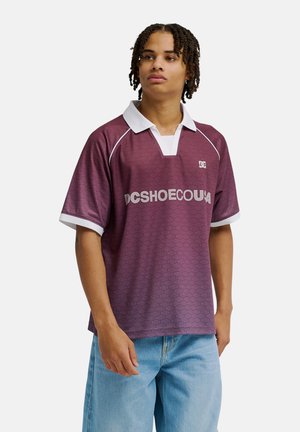 Jeune homme aux cheveux tressés, portant un polo bordeaux à manches courtes avec un col blanc et un jean bleu, debout devant un fond blanc uni.