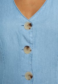 Jasnoniebieska sukienka dżinsowa z dekoltem w kształcie litery V, trzema okrągłymi, beżowymi guzikami z delikatnym wzorem oraz gładką, teksturowaną tkaniną.