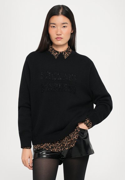 Svart strikket genser med "BRILLIANT" i teksturert detalj, lagd over en leopardmønstret skjorte og blanke svarte shorts.