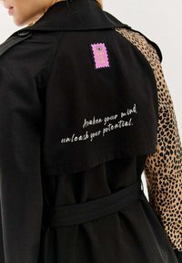 Čierny trenčkot s rukávom s leopardím vzorom, ktorý má ružovú nálepku a biele písmo s textom "Prebuď svoju myseľ, uvoľni svoj potenciál."