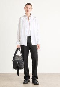 Camisa blanca de botones sobre una camiseta gris, combinada con pantalones negros. Bolsa negra con correa sostenida en una mano. Zapatos negros. Diseño minimalista.