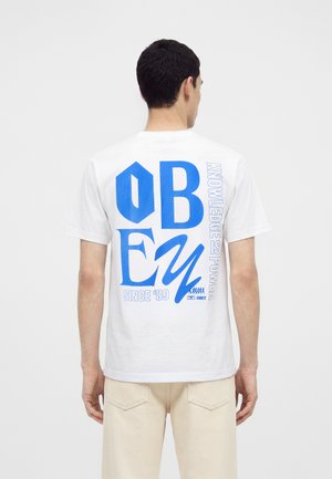 Muž v bielom tričku s veľkým modrým textom "OBEY" a "Knowledge is Power Since '89" na chrbte, stojaci pred jednotnou stenou.