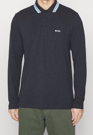 Man draagt een donkervlak polo met lange mouwen, met een blauw-witte gestreepte kraag en een "BOSS"-logo op de linkerborst, gecombineerd met een groene broek.