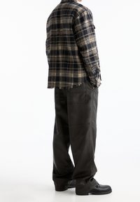 Personne portant une chemise à carreaux beige et noir avec des bords effilochés, un pantalon noir ample avec de grandes poches arrière, et des bottes noires.