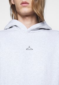 Felpa grigia chiara con coulisse, con un'icona di appendiabiti nera ricamata sul petto. Tessuto morbido, design casual.