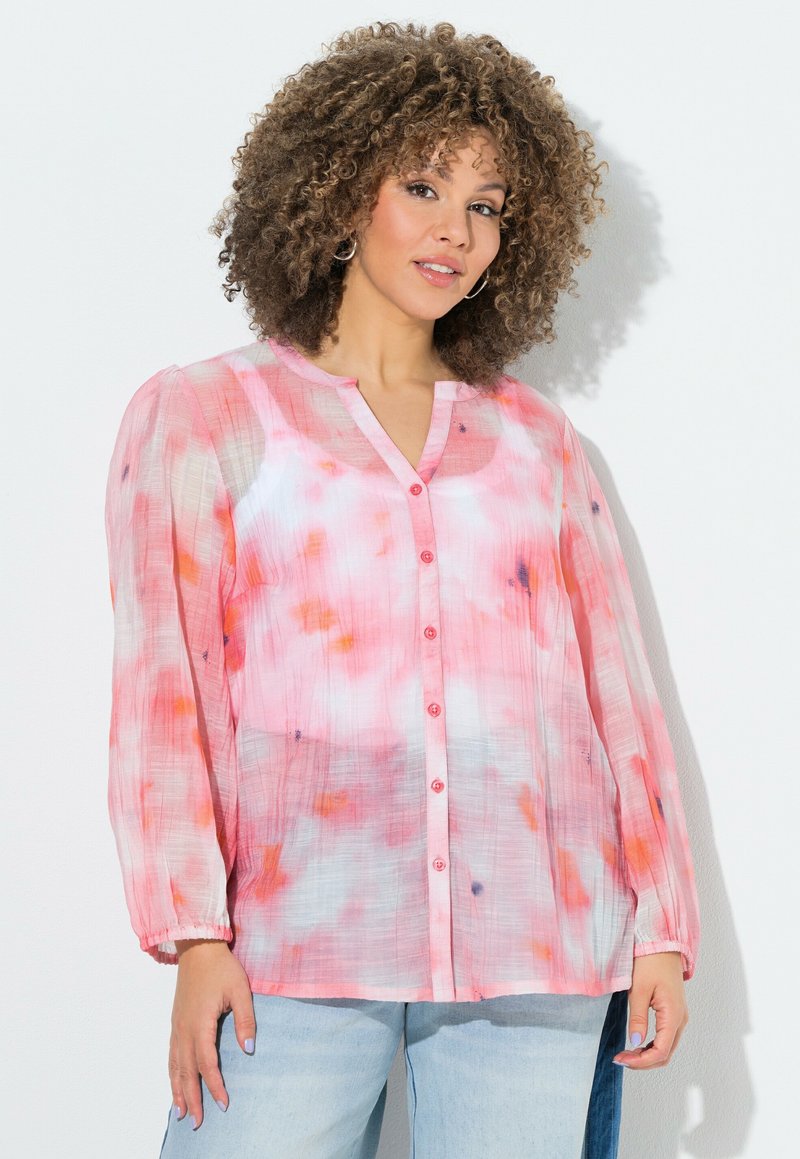 Kvinde med krøllet hår iført en pink, hvid og orange tie-dye skjorte med knapper og lange ærmer samt lyseblå jeans, stående foran en hvid baggrund.