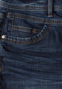 Jean en denim bleu foncé avec une poche avant présentant des coutures visibles et un rivet en argent. La texture est lisse avec un léger délavage.
