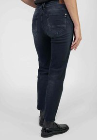 Jean en denim foncé avec une coupe slim, comportant deux poches arrière, une taille haute et des détails de couture minimaux. Porté avec des chaussures noires et des chaussettes grises.