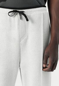 White textured jogging pants with a perforated pattern, featuring a black drawstring at the waist and side pockets.