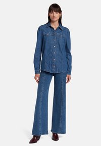 Denimjack met een puntige kraag, knoopsluiting en borstzakken. Gecombineerd met wijde denim broek, beide in medium blauw. Bruine accenten op de knopen.