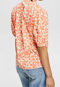 Kurzärmelige Bluse in Creme mit orangefarbenem Blättermuster, runder Halsauschnitt und gerafften Details an den Ärmeln. Tragen mit hellblauen Jeans.