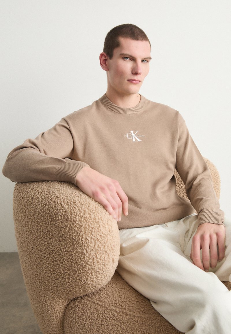 Μπεζ πλεκτό πουλόβερ με ριμπ λαιμόκοψη και μανίκια, διαθέτει λευκό λογότυπο στο στήθος. Το μοντέλο κάθεται σε μια υφασμάτινη, στρογγυλή καρέκλα.