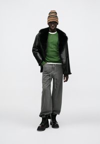 Μαύρο δερμάτινο μπουφάν με γούνα στο γιακά, πράσινο πλεκτό πουλόβερ, γκρι παντελόνι φαρδιάς γραμμής και μαύρα μποτάκια. Ριγέ πλεκτός σκούφος στο κεφάλι.