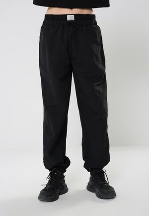 Personne portant un pantalon noir ample avec un cordon de serrage et des baskets noires, debout sur un sol gris avec un fond uni.