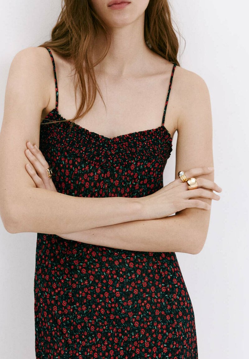 Femme aux longs cheveux bruns porte une robe noire sans manches avec un petit imprimé floral rouge, les bras croisés et des bagues en or aux doigts.