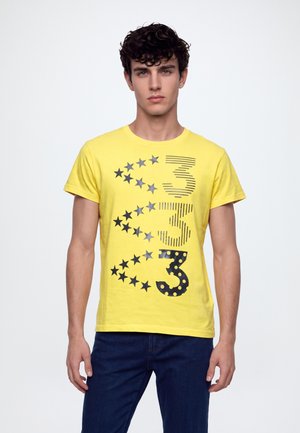 Un jeune homme porte un T-shirt jaune orné d’étoiles, de rayures et d’un grand chiffre 3, assorti à un jean foncé, sur un fond blanc uni.