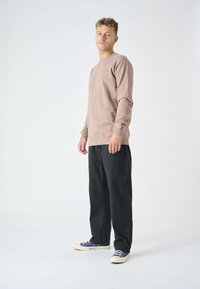 Light brown long-sleeve shirt with a round neckline, worn with loose black pants and blue sneakers. Simple design, soft texture, minimal detailing.