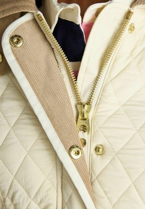 Kreemjas tikitud jakk beežist sametvoodriga, kuldse tõmbluku ja neetidega, osaliselt lahtiläinud, et näidata allolevat triibulist särki.