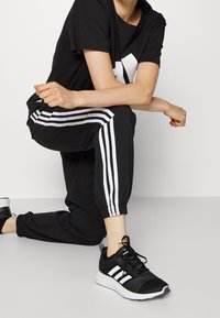 Czarny strój sportowy składający się z t-shirtu z dużym białym logo oraz czarnych spodni dresowych z białymi paskami po bokach. Czarne buty sportowe z białymi elementami.