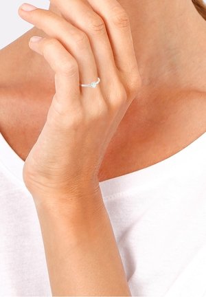 Main à la peau claire portant une bague en or blanc avec un diamant solitaire à l'annulaire, reposant près de la poitrine dans un haut blanc.