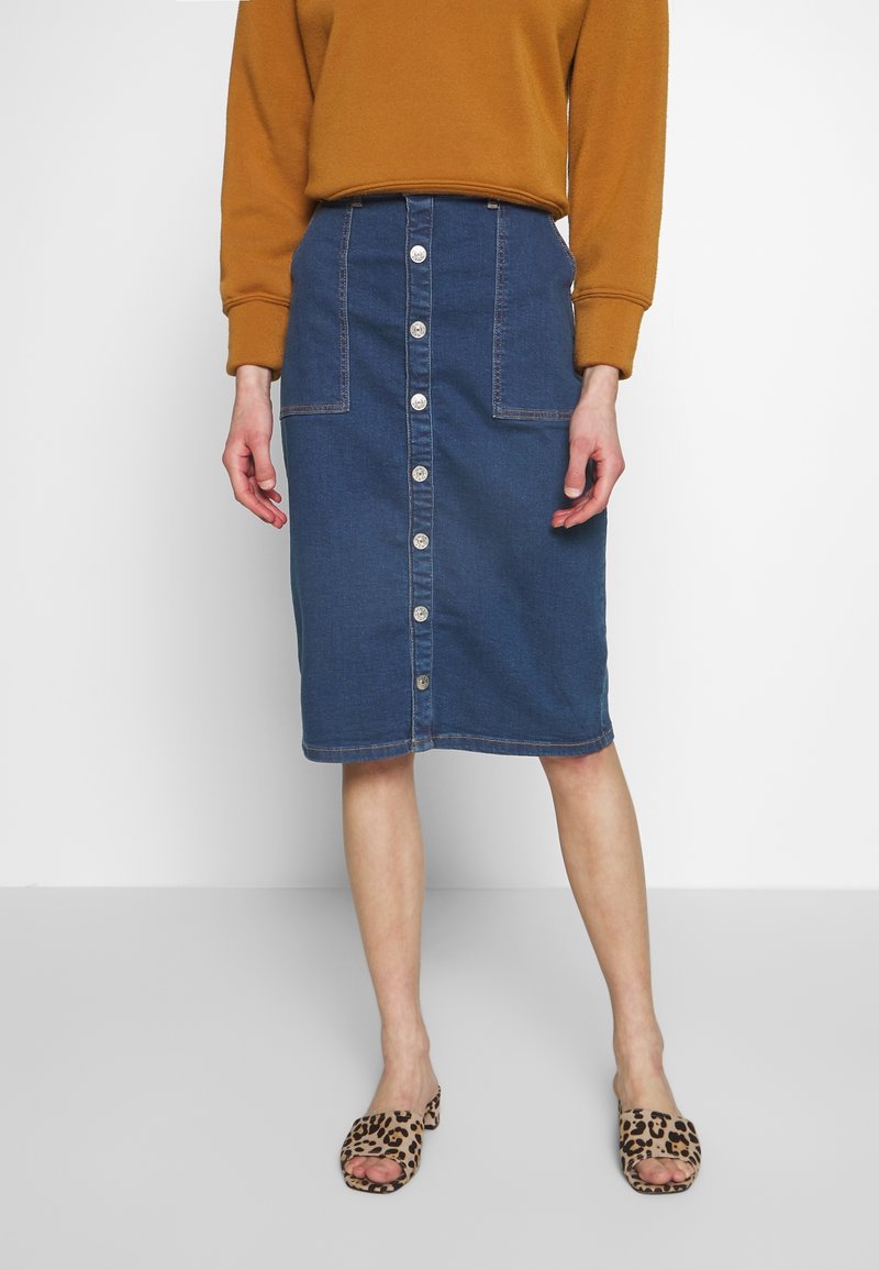 Denim A-line skirt in dark blue with front button closure and pockets. Paired with a brown sweatshirt and leopard print slides.