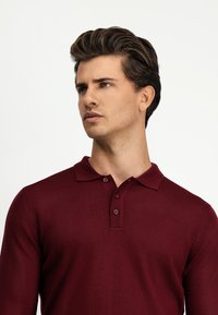 Burgundowa polo w kolorze czerwonym z długim rękawem o fakturze żebrowej, z trzema guzikami i płaskim kołnierzykiem, z delikatnymi detalami wzdłuż szwów.