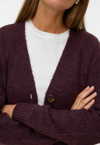 Gilet en tricot torsadé bordeaux avec des manches longues, un décolleté en V profond et une fermeture à un bouton rond. Porté sur un t-shirt blanc. Texture douce.