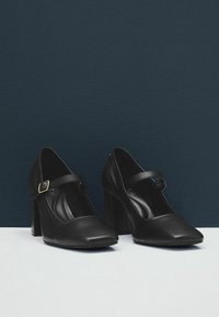 Černé kožené baleríny Mary Jane s čtveratými špičkami a chunky blokovými podpatky, vystavené na bílé ploše na pozadí tmavě modré barvy.