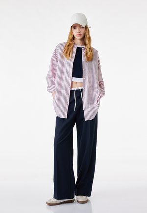 Joven mujer con gorra blanca, camisa rosa de rayas oversize, top corto negro, pantalones anchos negros y zapatillas blancas, de pie con las manos en los bolsillos.