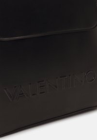 Černý kožený batoh s vyraženým logem "VALENTINO", hladkou texturou, klopou na zavírání a čistým, minimalistickým designem.