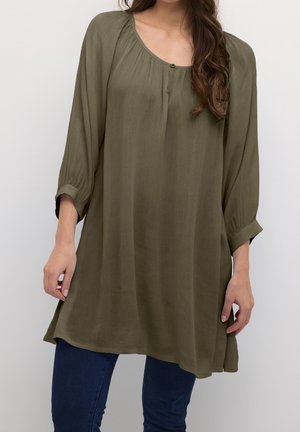 Frau trägt eine locker sitzende, olivgrüne Langarm-Tunika mit geknöpftem Ausschnitt, kombiniert mit dunkelblauen Jeans vor einem schlichten hellen Hintergrund.