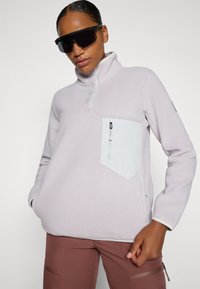 Heled lilla fliisipullover, millel on poolne lukustus, valge rinnatasku ja küljetaskud. Omab kõrget kaelust ja kontrastset äärestust.