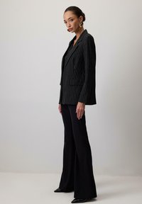 Sacou negru cu dungi, ajustat, cu rever în formă de v, și închidere cu un singur nasture, purtat cu pantaloni negri evazați și pantofi ascuțiți.