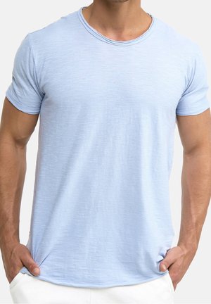 Man wearing light blue short-sleeve crew neck t-shirt with hands in pockets against white background.
