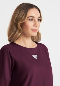 Bordeaux lange mouw top met ronde hals; heeft een witte hartontwerp met het woord "FREEDA" op de borst. Zijdezachte stoffen textuur.