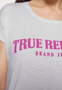 Bílý tričko s krátkým rukávem a kulatým výstřihem, na kterém je vynikající růžový text "TRUE RELAX" a "BRAND JEANS" potisknutý na přední straně.