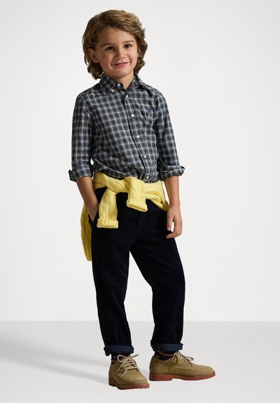 Camisa de cuadros abotonada en tonos oscuros, suéter amarillo atado a la cintura, pantalones de pana oscuros y zapatos de cordones color caqui.