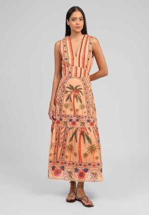 Femme debout portant une robe sans manches, col en V, longueur jusqu'aux chevilles avec des imprimés de palmiers et de fleurs, associée à des sandales plates à brides.